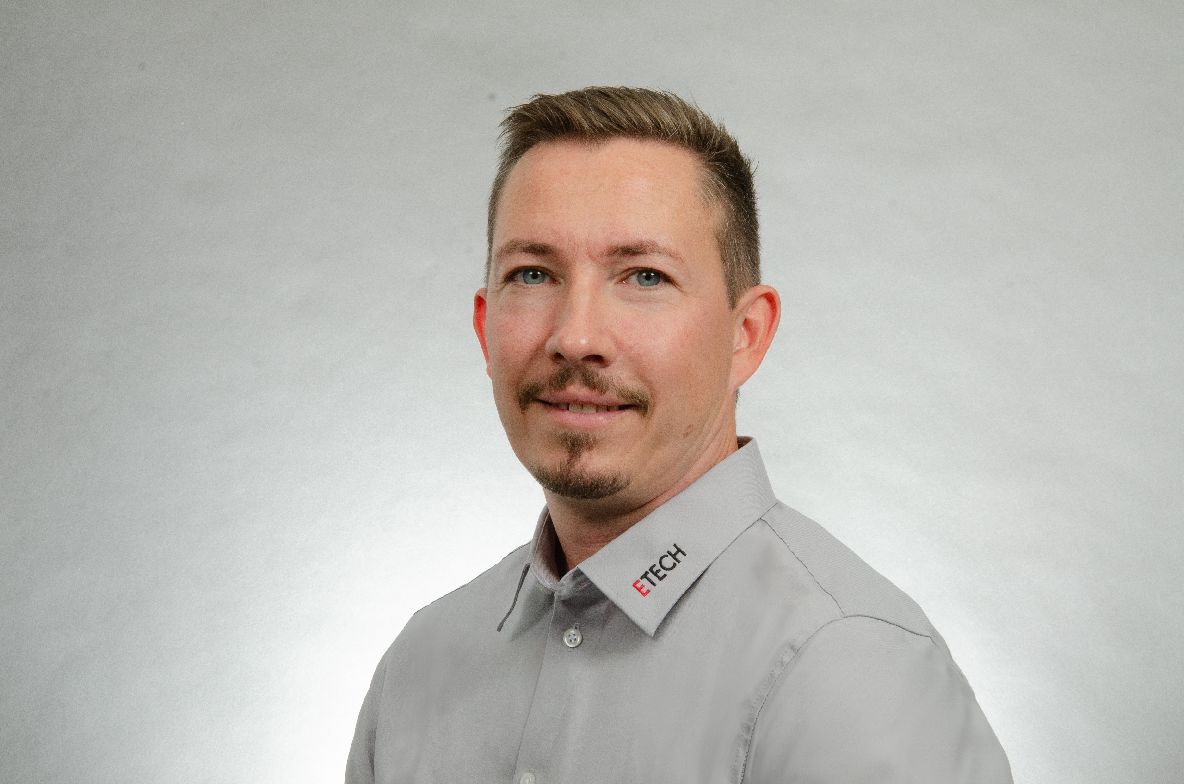 Mayr Christian ETECH Mitarbeiter Porträt eines lächelnden Mannes in grauem Hemd mit ETECH-Logo auf dem Kragen vor neutralem Hintergrund.